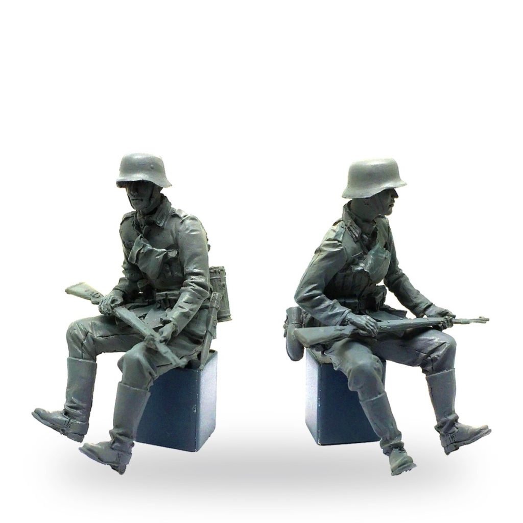 Two unpainted WWII German soldier figures sitting with rifles on boxes.